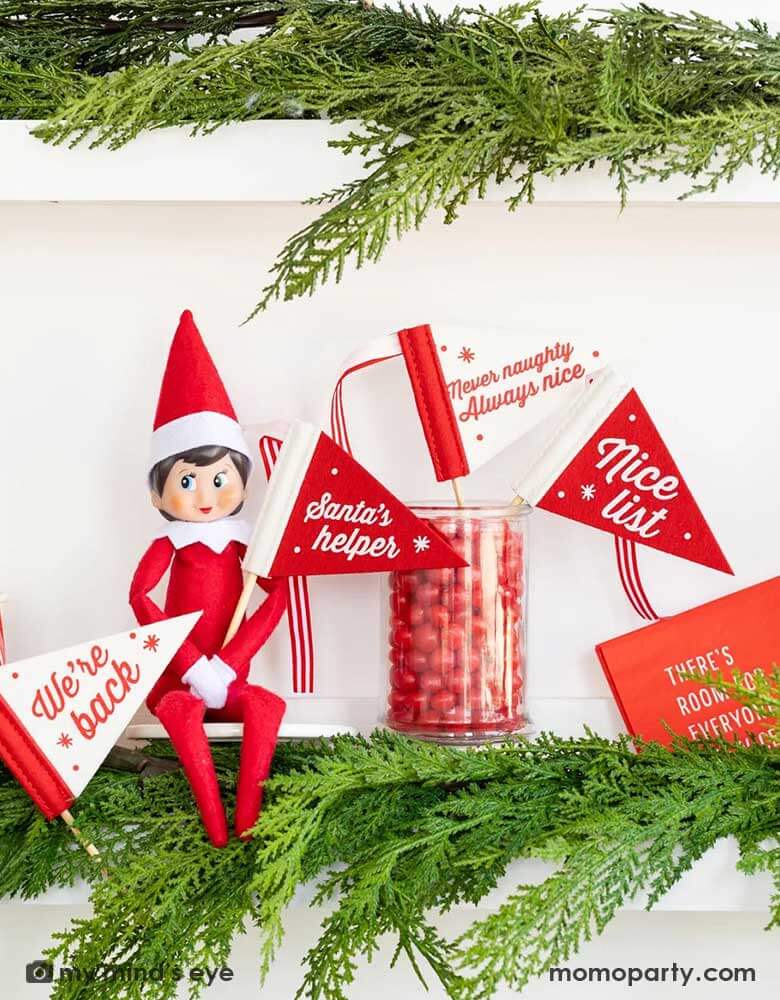 An elf on the shelf sitting on a white shelf decorated with green Christmas tree branches and leaves, on his hands held a red and white Momopartys's Christmas Elves on the Shelf felt pennant banners flag by My Mind's Eye featuring words "Santa's Helper", along with other felt banner flags featuring "We're back," "Never Naughty Always Nice", and "Nice List," along with some red jelly beans in a candy jar. All makes a great idea for kid's Holiday elf on the shelf Christmas home activity.