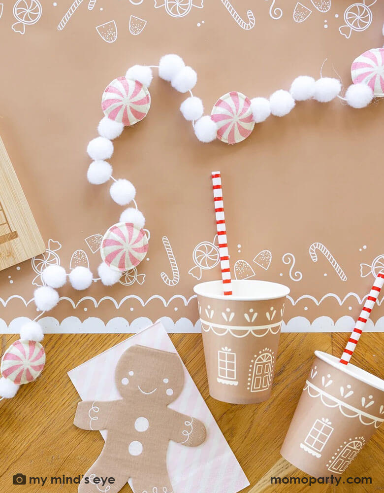 A tabletop featuring Momopartys's gingerbread man table runner, on top of it are the 4.25" x 7.75" gingerbread man shaped napkins by My Mind's Eye, along with a bamboo gingerbread house shaped serving tray which has two cute gingerbread man cookies on tis, next to it lay a pink candy cane felt banner which is surrounded with two gingerbread house party cups and pink striped dinner napkins from Momopartys for a kid's cookie decorating / gingerbread house building party.