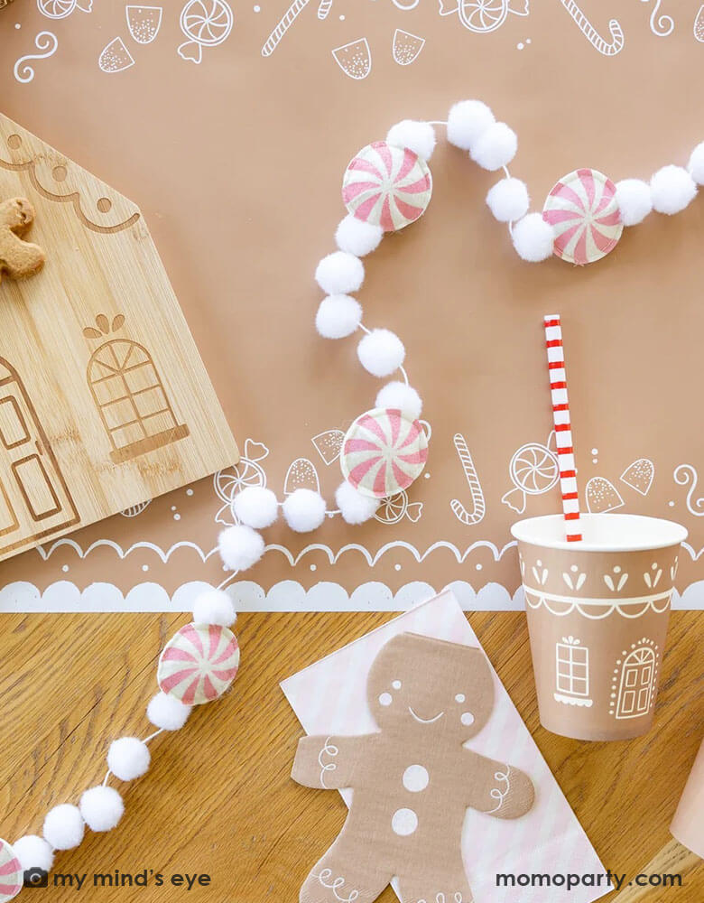 A tabletop featuring Momopartys's gingerbread man table runner, on top of it are the 4.25" x 7.75" gingerbread man shaped napkins by My Mind's Eye, along with a bamboo gingerbread house shaped serving tray which has two cute gingerbread man cookies on tis, next to it lay a pink candy cane felt banner which is surrounded with two gingerbread house party cups and pink striped dinner napkins from Momopartys for a kid's cookie decorating / gingerbread house building party.