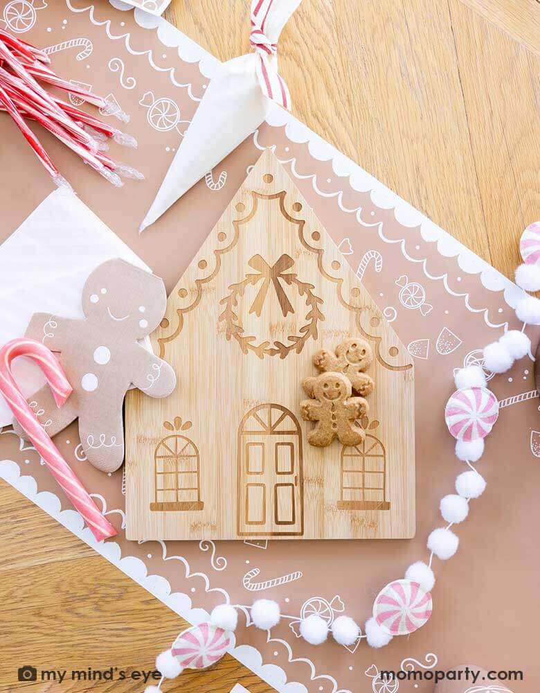 A tabletop featuring Momopartys's gingerbread man table runner, on top of it are the 4.25" x 7.75" gingerbread man shaped napkins by My Mind's Eye, along with a bamboo gingerbread house shaped serving tray which has two cute gingerbread man cookies on tis, next to it lay a pink candy cane felt banner which is surrounded with candy canes and cookie decoration butter cream piping bags for a kid's cookie decorating / gingerbread house building party.