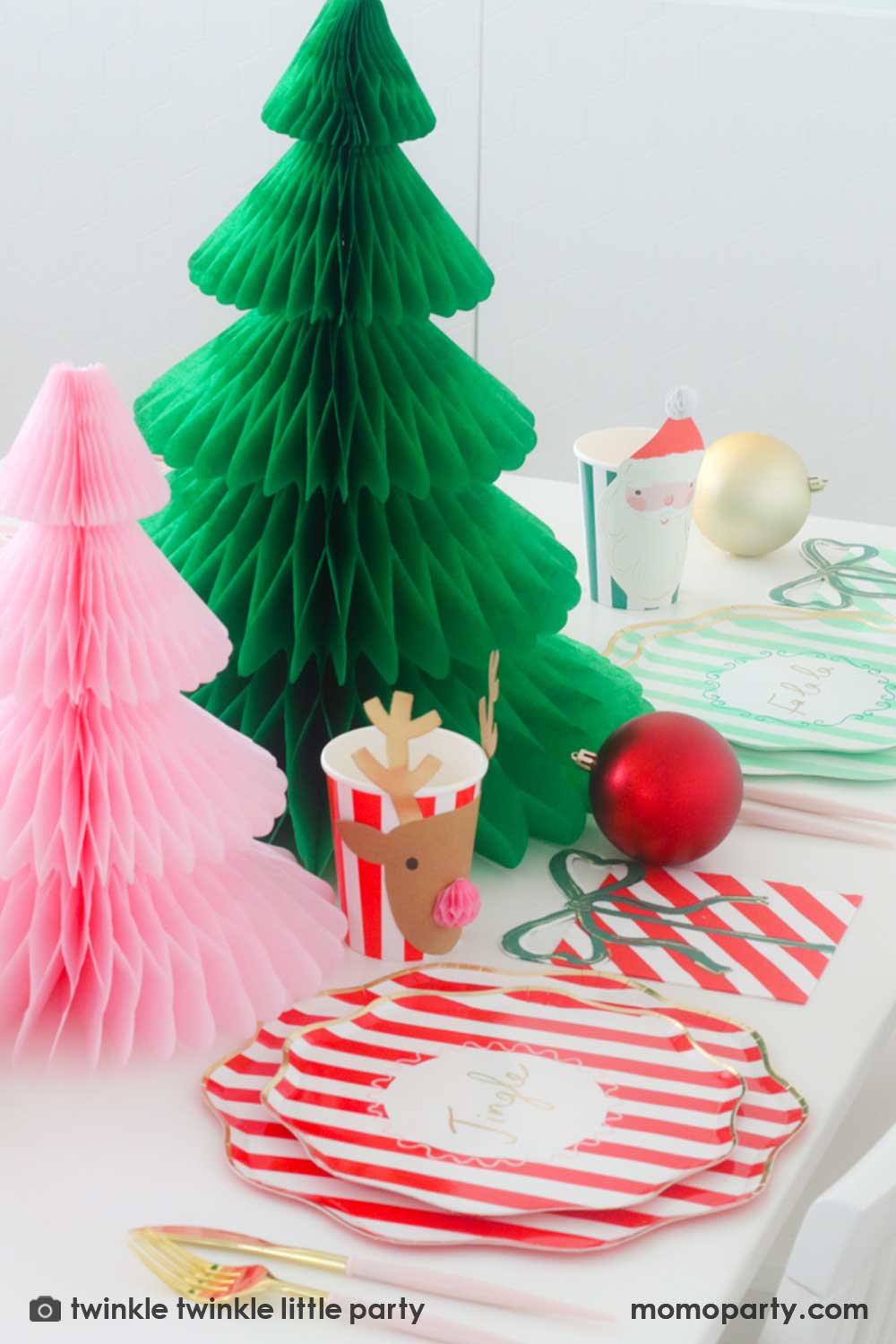 A festive Christmas party table featuring Momopartys's Festive striped dinner plates, side plates, present with a bow shaped napkins and Christmas honeycomb party cups in Santa design by Meri Meri. In the center of the table are a green and pink honeycomb Christmas tree decorations as the centerpiece. Along with some Christmas ornaments on the table, this makes a modern Holiday place setting inspo for a festive party this holiday season.