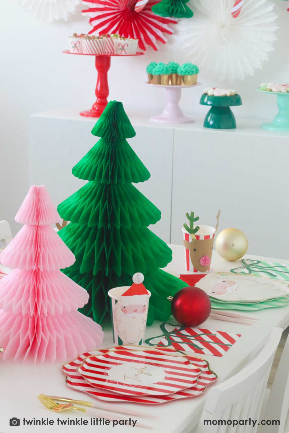 A festive Christmas party table featuring Momopartys's Festive striped dinner plates, side plates, present with a bow shaped napkins and Christmas honeycomb party cups in Santa design by Meri Meri. In the center of the table are a green and pink honeycomb Christmas tree decorations as the centerpiece. Along with some Christmas ornaments on the table, this makes a modern Holiday place setting inspo for a festive party this holiday season.