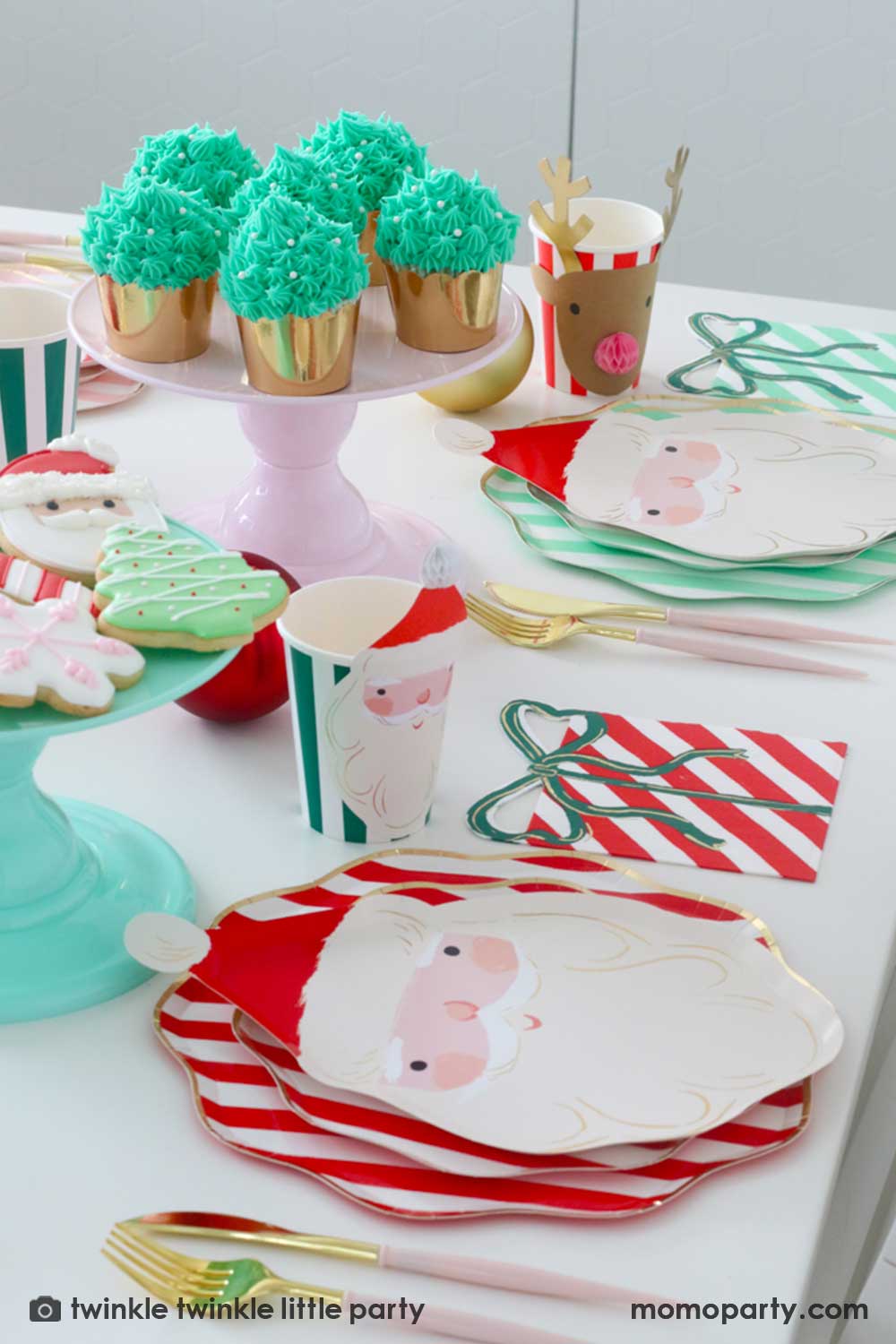 A festive Christmas party table featuring Momopartys's Festive striped dinner plates, side plates, Jolly Santa Plates, present with a bow napkins and Christmas honeycomb party cups by Meri Meri. On the center of the table are some buttercream Christmas tree cupcakes in festive gold foil baking cups and a mint cake topper with festive Holiday cookies in the design of a Santa, Christmas tree, candy cane and snowflake. Makes this a modern inspo for a festive Holiday table setting.