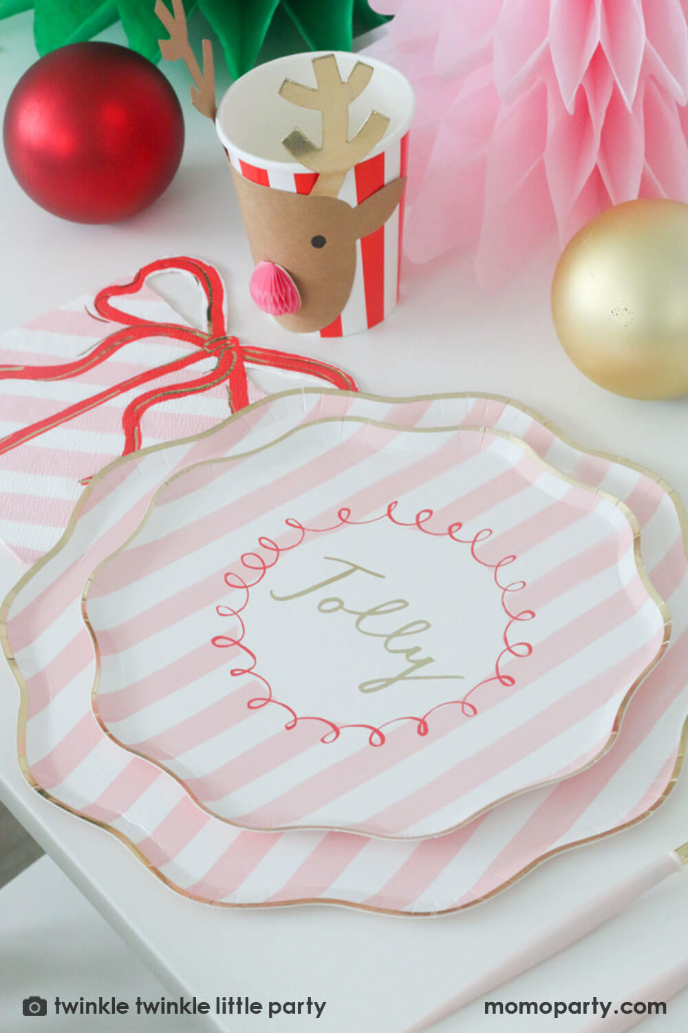 A festive Christmas party table featuring Momopartys's Festive pink striped dinner plate layered by a side plate and a pink present with a bow shaped napkin and a reindeer red honeycomb party cup by Meri Meri. In the center of the table are a green and pink honeycomb Christmas trees along with red and gold Christmas ornaments, making this a gorgeous Holiday tablescape for a darling Christmas party this holiday season.