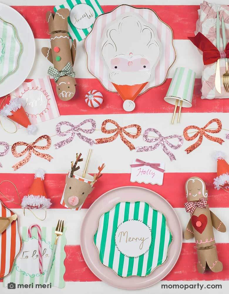 A festive Christmas party table featuring Momopartys's new Christmas party collection including festive striped plates, napkins and party cups in green, pink and red. On the red striped tablecover, there are also Meri Meri's Christmas honeycomb party cups in reindeer design, which paired with Santa shaped plates and glittering red and pink bows garland in the middle, makes it a festive and fun tablescape for kid's friendly Christmas party.
