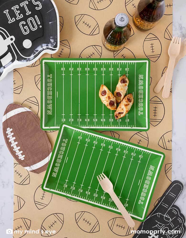 A football party table features Momopartys's Football Field shaped plates, on the plate there are some party snacks and around the plates there are Momopartys's football shaped napkins, black helmet shaped plates featuring "Let's Go"" on it, and a no #1 finger shaped black napkin on a light brown table runner with football illustration on it. With party drinks around, it makes a great party table decoration for a fun football season watch game party or a Super Bowl party!