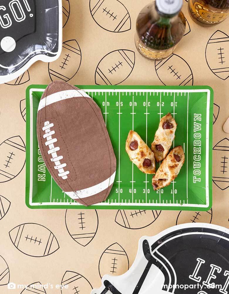 A football party table features Momopartys's Football Field shaped plate, on the plate there are some party snacks and some football shaped napkins, around the plates there are Momopartys's football shaped black helmet shaped plates featuring "Let's Go"" on them. All these are on a light brown table runner with football illustrations on it. With party drinks around, it makes a great party table decoration for a fun football season watch game party or a Super Bowl party!