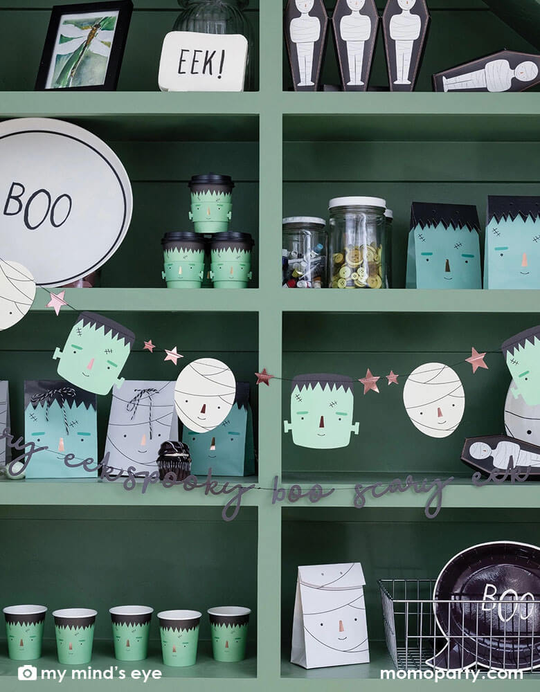 A green shelf filled with Momopartys's Halloween party supplies including Frankenstein and mummy themed plates, cups, goodie bags, treat boxes and napkins. In the front of the shelf, there's a Frank & Mummy garland hung, featuring spooky best friends, copper foiled stars, and a stitched word banner, these banners a sure to be a frightful delight at your Halloween parties!