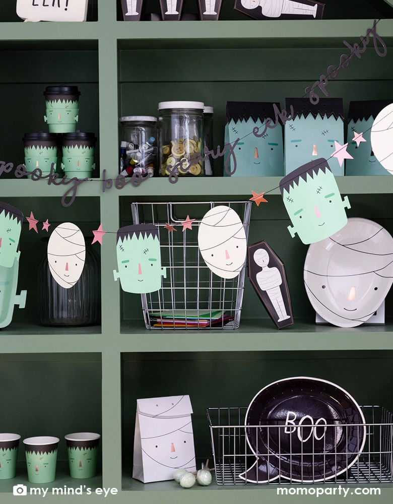 A green display shelf filled with Momopartys's Halloween party supplies including Frankenstein and mummy themed plates, cups, goodie bags, treat boxes and napkins. In the front of the shelf, there's a Frank & Mummy garland hung,  featuring spooky best friends, copper foiled stars, and a stitched word banner, these banners a sure to be a frightful delight at your Halloween parties!