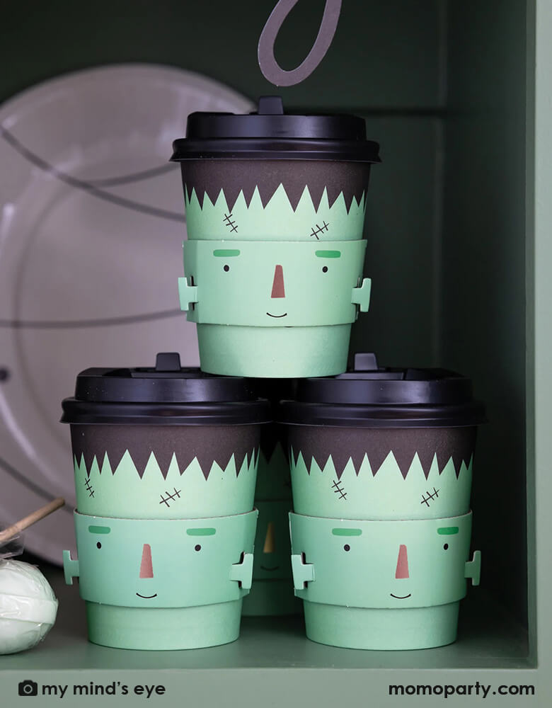Three Momopartys's 8 oz Frank and Mummy Cozy to-go cups by My Mind's Eye stacked together on a shelf. Featuring a friendly Frankenstein sleeve these cozy to go cups will keep your favorite beverage warm through all the tricks and treats on Halloween night!