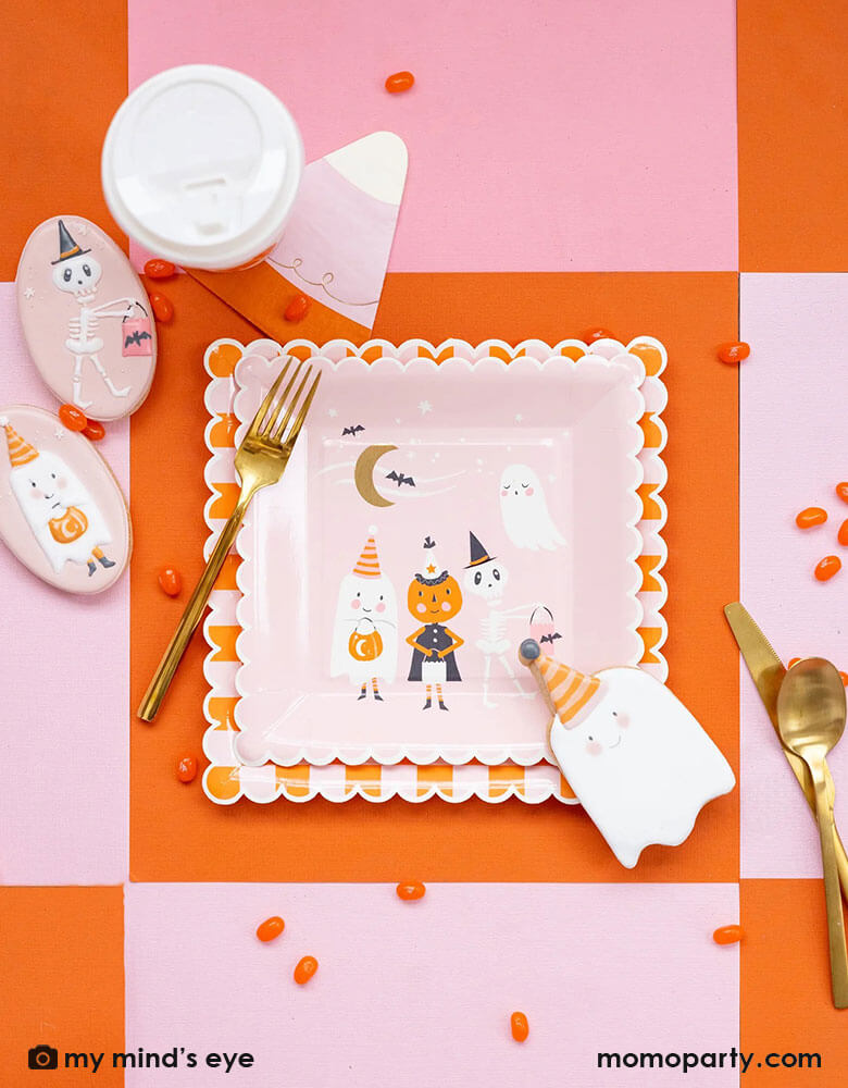 A pink Halloween party table with orange and pink checkered tablecover, featuring Momopartys's ghoul gang collection by My Mind's Eye including ghoul gang scene scallop edged square plates and orange and pink checkered scallop edged plates. With coordinating decorated Halloween sugar cookies in the adorable colors of light pink and orange with orange jelly beans around the table, it makes an adorable pink girly Halloween party inspiration.