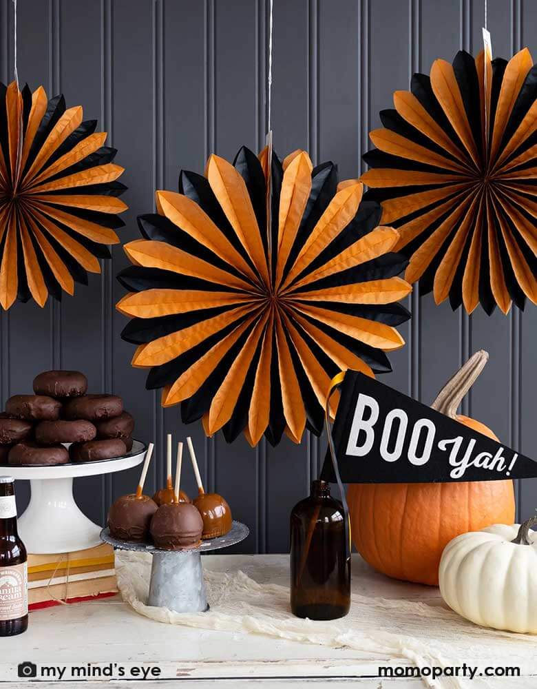 A classic orange and black Halloween table decorated with pumpkins, caramel apples and Momopartys's 14" x 15" Boo Yah black and white felt party pennant with orange and black ribbons. In the back the wall was decorated with My Mind's Eye's oversized orange and black tissue paper fans. All makes a simply yet iconic Halloween decorating inspo this Halloween season.