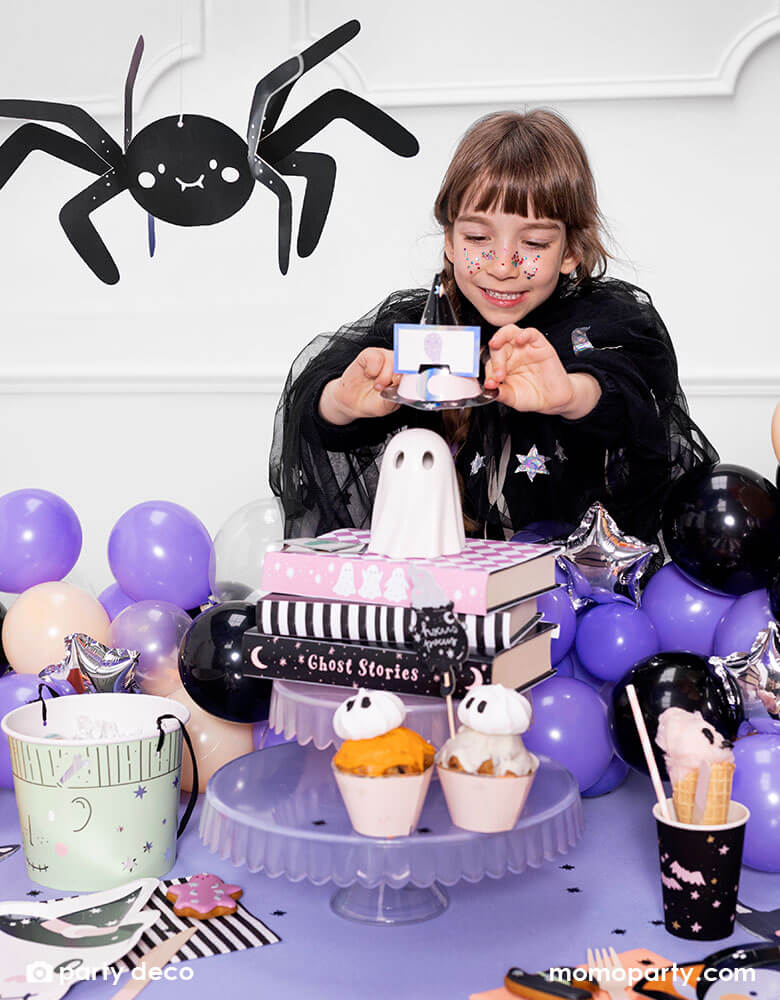 A girl playing with Momopartys's Halloween Guessing Game Set by Party Deco at a Halloween party table decorated with a purple tablecloth and black balloons. On the table there are some Halloween themed treats including ghost cupcakes, pink cotton candy, Halloween sugar cookies on Momopartys's pink skull shaped plates. Above the party table there's an adorable smiley spider paper decoration hung above, makes it a cute inspiration for kid-friendly Halloween party set up.