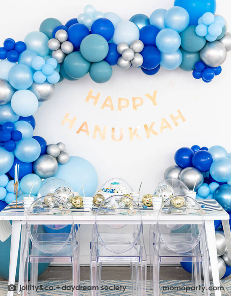 A festive Hanukkah party table set up featuring a large blue balloon garland and "Happy Hanukkah" letter banner above the party table. On the table are Momopartys's blue striped plates and party cups with Festival of Lights small star shaped plates and patterned napkins by Daydream Society. On the blue striped table runner are some silver and gold disco balls in different sizes and Chanukah candles. All makes it a great inspo for a festive Hanukkah party for family gathering.