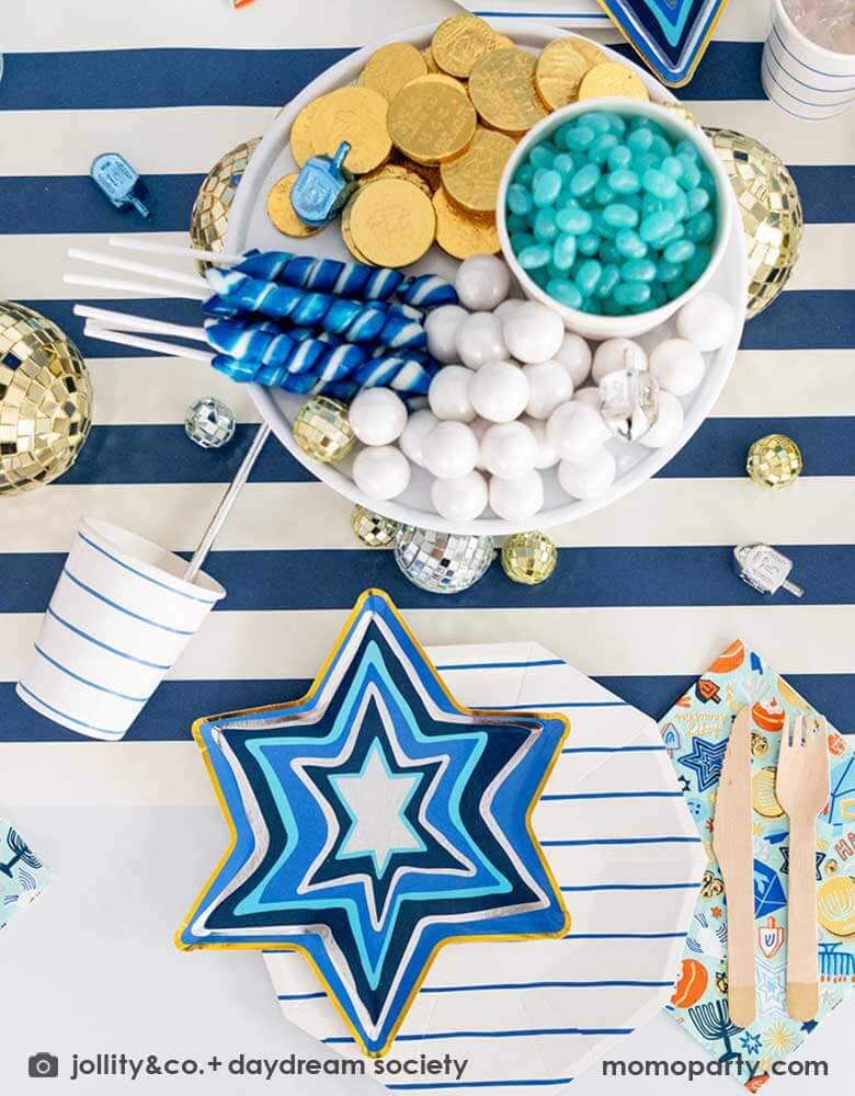 A festive Hanukkah party table featuring Momopartys's blue striped plates and party cups with Festival of Lights small star shaped plates and patterned napkins by Daydream Society. On the a blue striped table runner is a cupcake holder with treats and candies in coordinating blue and white colors and some coin chocolates and Dreidel candies, makes it a great inspo for a festive Hanukkah party for family gathering. 