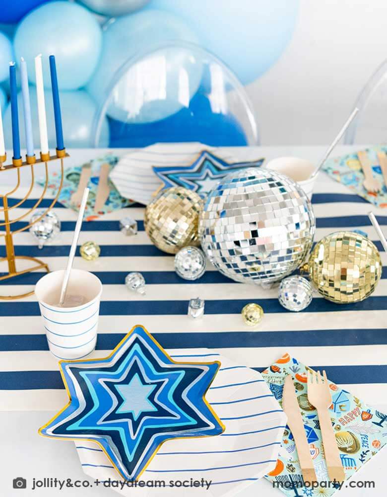 A festive Hanukkah party table featuring Momopartys's blue striped plates and party cups with Festival of Lights small star shaped plates and patterned napkins by Daydream Society. On the blue striped table runner are some silver and gold disco balls in different sizes and Chanukah candles. In the back you can see a blue balloon garland in different shades of blue, makes it a great inspo for a festive Hanukkah party for family gathering.