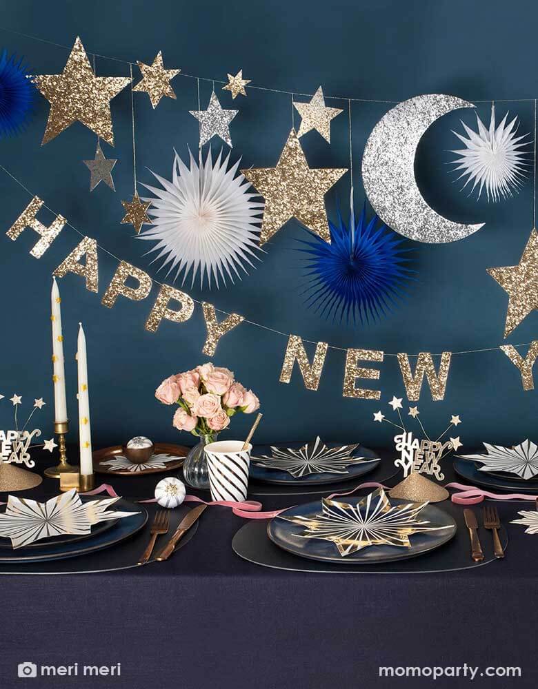 A festive New Year Party Table featuring Momopartys's gold star striped plates and napkins, gold striped party cups and Happy New Year party hats on a dark navy covered table. Above the table hung Momopartys's 8 ft New Year party garland with gold and silver glittered moon and star pennants, along with navy and white paper fans, below it is a gold glittered Happy New Year letter banner, makes it a great inspiration for a festive yet elegant New Year countdown party.