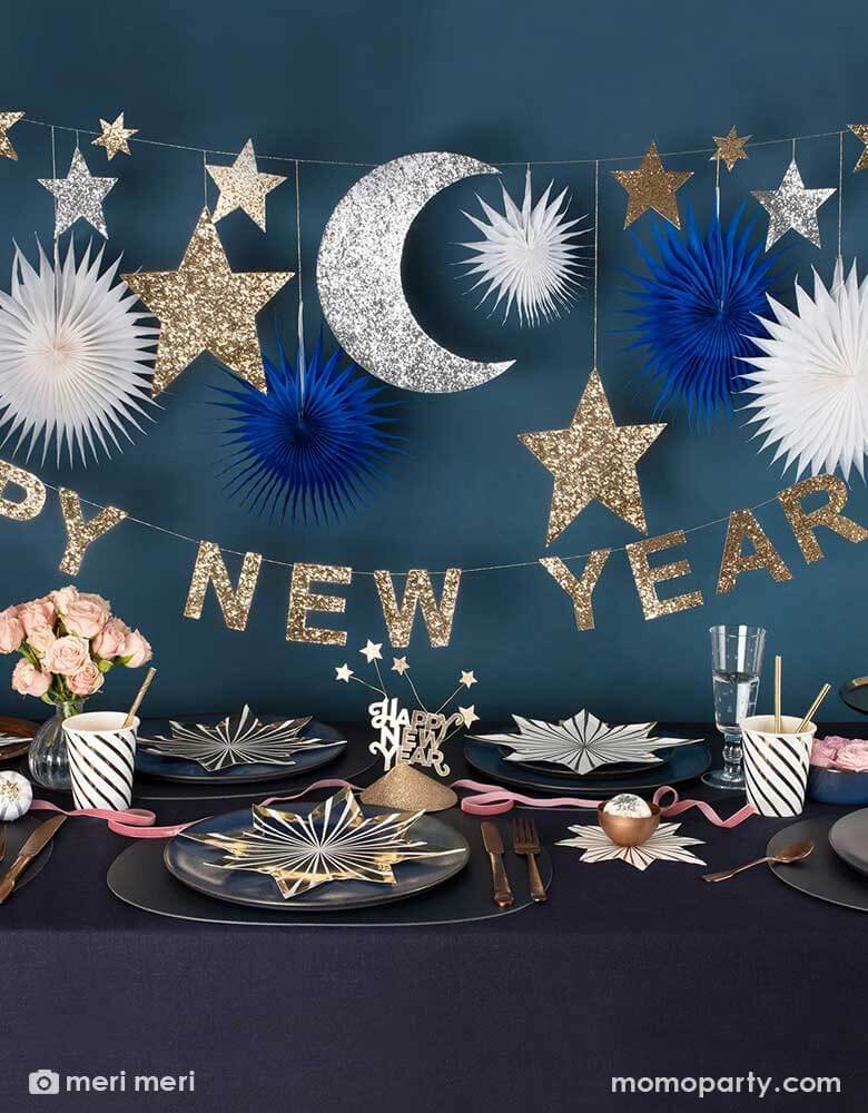 A festive New Year Party Table featuring Momopartys's gold star striped plates and napkins, gold striped party cups and Happy New Year party hats on a dark navy covered table. Above the table hung Momopartys's 8 ft New Year party garland with gold and silver glittered moon and star pennants, along with navy and white paper fans, below it is a gold glittered Happy New Year letter banner, makes it a great inspiration for a festive yet elegant New Year countdown party.