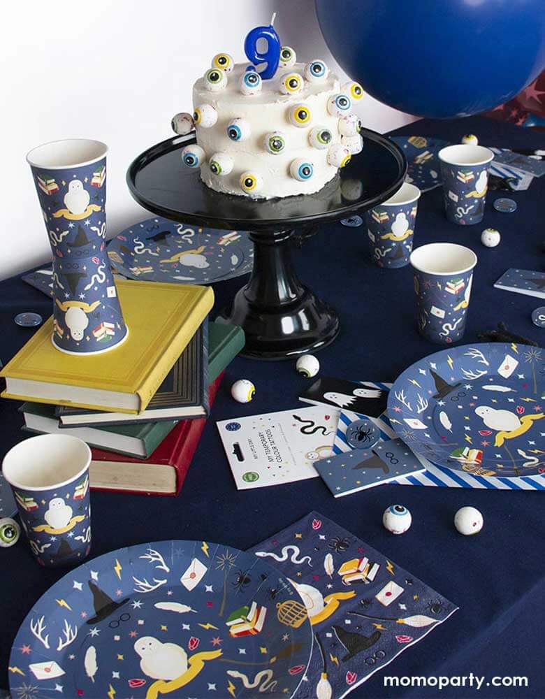A Harry Potter wizard themed party table featuring Momopartys's wizard party supplies including party plates, cups, napkins and temporary tattoos. With a cake decorated with spooky eyeballs as the centerpiece and spell books in the middle, this makes a great inspiration for kid's Harry Potter themed birthday party or Halloween bash!