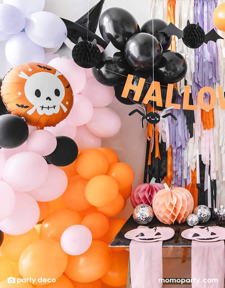 A festive Halloween party set up featuring a beautiful balloon garland in orange, pink, lilac and black, with Momopartys's 14" orange round skull foil balloon, witch's hat, paper spider hanging decoration and honeycomb bats in front of a wall of colorful fringe in the Halloween themed colors. In the front there's a party table with some honeycomb pumpkins, disco balls and pink pumpkin shaped plates. All makes this a perfect party inspiration for a not so spooky kid's friendly Halloween party.