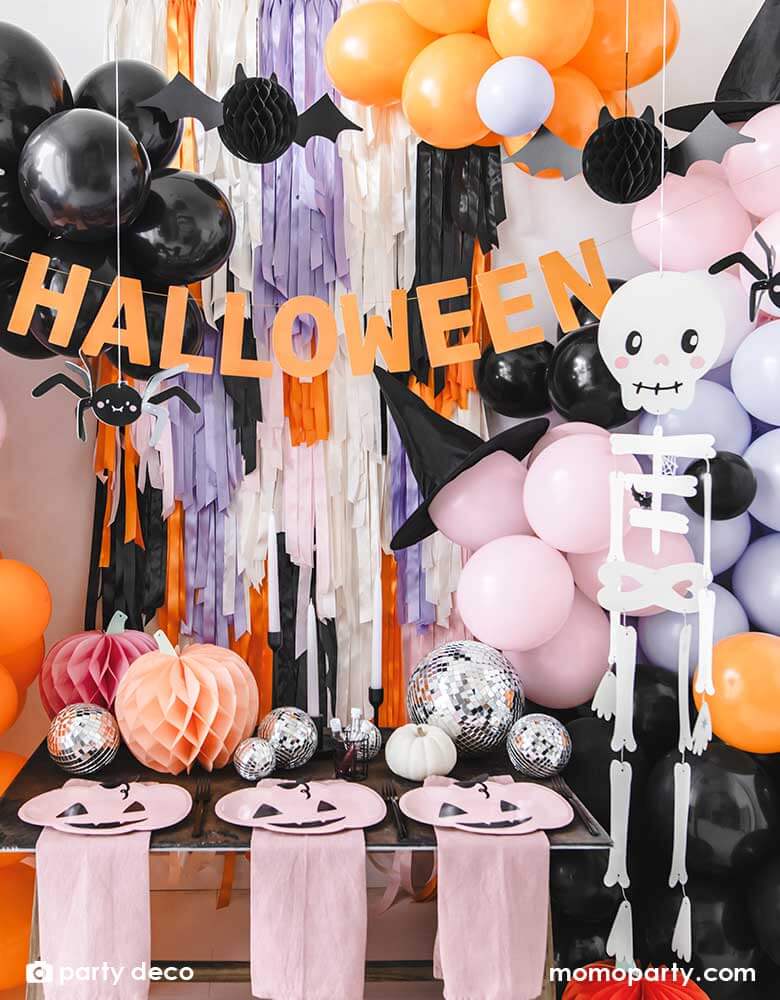 A festive Halloween party set up featuring a beautiful balloon garland in orange, pink, lilac and black, with Momopartys's 14" orange round skull foil balloon, witch's hats, skeleton hanging decoration, paper spider hanging decoration and honeycomb bats in front of a wall of colorful fringe in the Halloween themed colors. In the front there's a party table with some honeycomb pumpkins, disco balls and pink pumpkin shaped plates. 