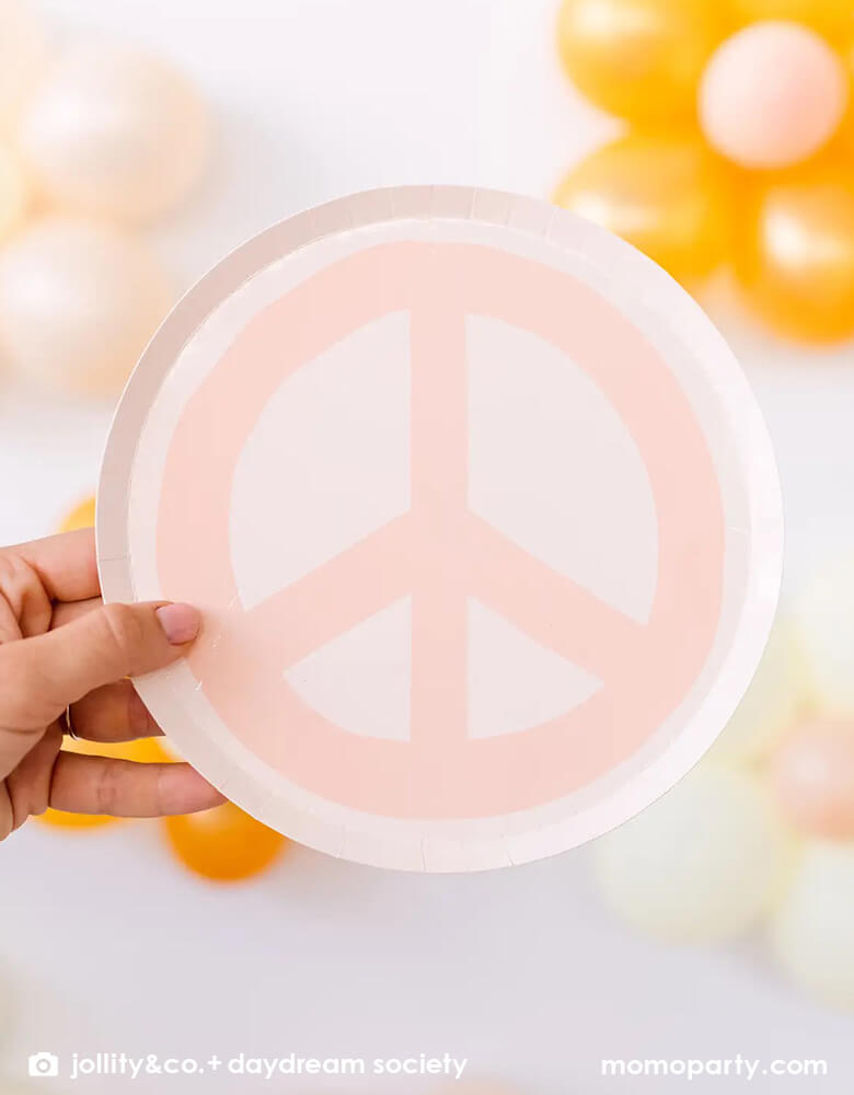 A hand holding Momopartys's 8" peach love and peace sign dessert plate by Jollity & Co. in front of a wall decorated with daisy balloons. Momopartys's 8" love and peace smily face dessert plate by Jollity & Co. Featuring a groovy, retro design and bursting with boho vibes, these plates will bring good vibes to any girl's birthday.