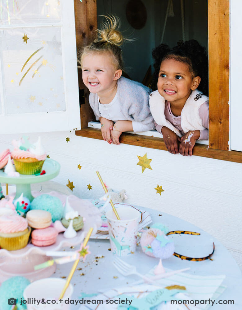 Two school aged girls leaning against a window looking out a party table featuring Momopartys's magical unicorn partyware including plates and cups. In the middle of the table, a cupcake tower with a sorts of treats including cupcakes, macarons, cotton candies and more, makes this a whimsical Christmas holiday set up that's perfect for kids.