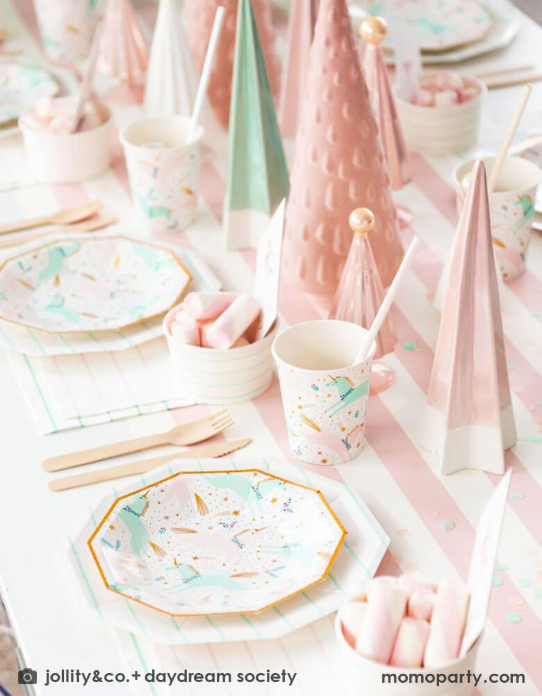 A pink Christmas party table featuring Momopartys's magical unicorn plates and cups by Daydream Society. A few ceramic Christmas tree decorations in mint and pink are used as the centerpiece on a pink white striped table runner. On the table you can see some pink marsh mellows in food cups as the treats for kids.