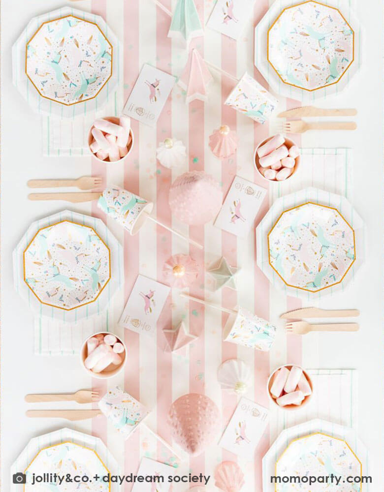 A pink Christmas party table featuring Momopartys's magical unicorn plates and cups by Daydream Society. A few ceramic Christmas tree decorations in mint and pink are used as the centerpiece on a pink white striped table runner. On the table you can see some pink marsh mellows in food cups as the treats for kids. All makes this a dreamy whimsical kid's holiday party inspo. 