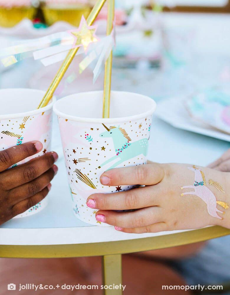 Two girl's hands holding Momopartys's magical unicorn party cups by Daydream Society. On on of the girls' hand there's a temporary tattoo of a unicorn on it. In the party cups, there are gold foil straws decorated with shooting star toppers to create a magical and whimsical vibe for a Christmas Holiday celebration party.