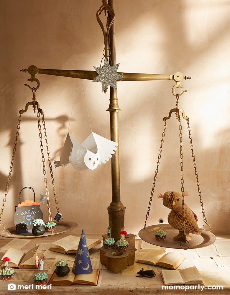 A vintage scale decorated with Momopartys's Making Magic party supplies collection by Meri Meri including honeycomb owls in brown and black, cauldron shaped party bags, witch hats and witch themed cupcake toppers. On the table are some spell books to add a little eerie and whimsical vibe to a wizard & witch themed party of a Harry Potter themed celebration.