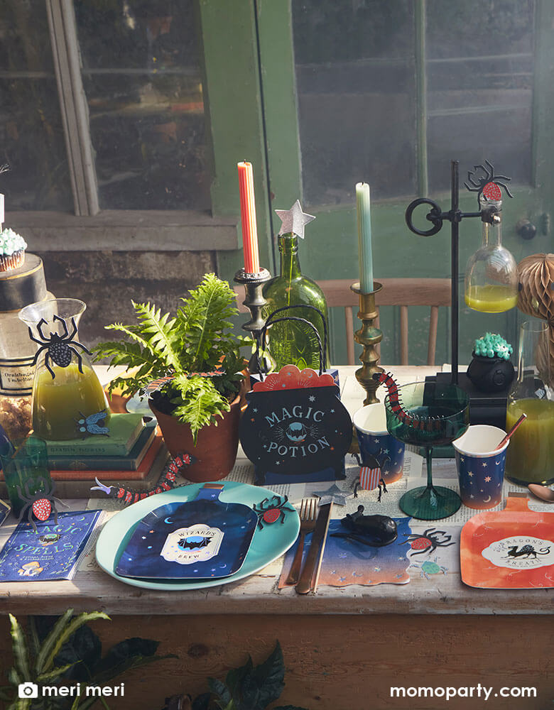 A witch and wizard party table set up featuring Momopartys's making magic party supplies collection by Meri Meri including potion bottle shaped plates, wizard spell book napkins, witch cauldron shaped party bags, owl honeycomb decoration, night ombre napkins and cups with moon and stars on them, and bug confetti scattered on the table, makes it a great inspiration for kid's harry potter themed party or a Halloween witch and wizard themed bash.