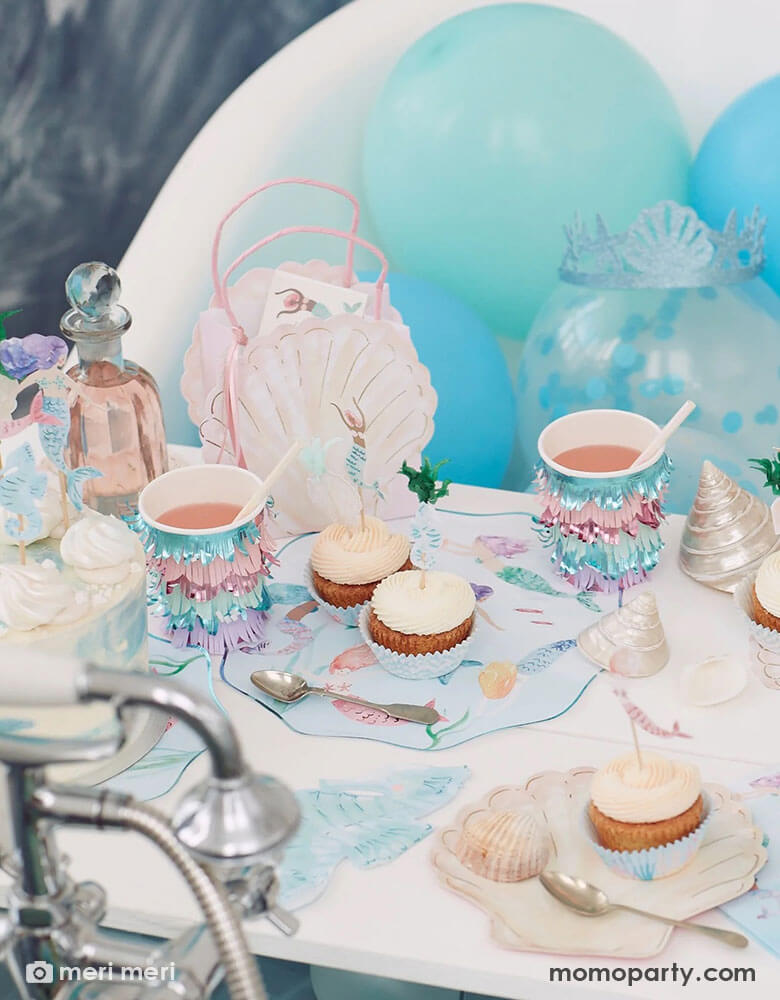 A beautiful kid's under the sea mermaid themed party table featuring Momopartys's mermaid party supplies including plates, iridescent fringe party cups, shell shaped plates, seahorse shaped napkins and pink shell shaped party bags by Meri Meri. In the back there are some fun latex balloons in light blue shades. On the table there are some cupcakes topped with mermaid themed cupcake toppers, makes this a great inspo for a whimsy mermaid party for girls.