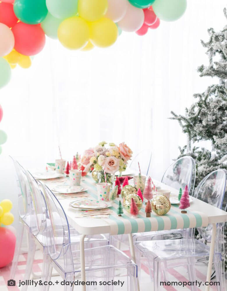 A Christmas table featuring Momopartys's Merry and Bright party collection by Daydream Society. On the table you can see the festive Christmas themed plate, cups and napkins on a mint striped table runner. The middle of the table is decorated with bottle brush Christmas trees in pastel colors. Above the party table, there is a balloon garland in the colors of coral, yellow, pink and mint hung over it. Finally on the right of the table, there's a big Christmas tree covered with frost.