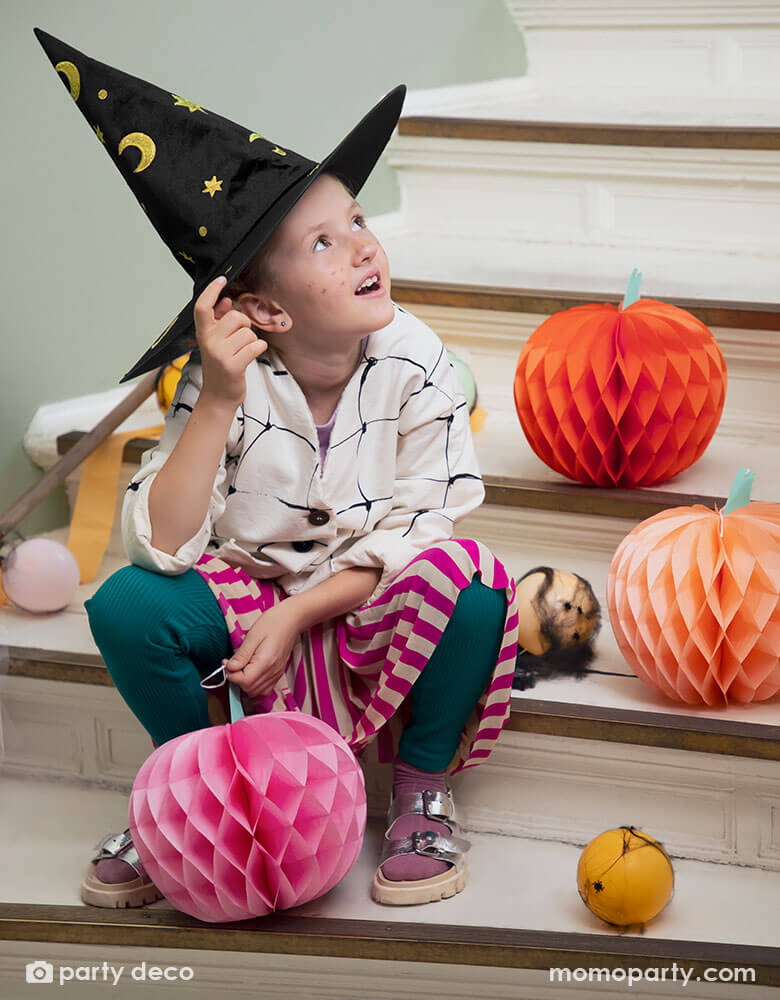 A girl wearing her Halloween costume and a witch hat setting on stairs looking up. Around her there are some party balloons in Halloween colors of orange and pink and blush wrapped in spider web to make a spooky vibe, along with Momopartys's 9.5" pink pumpkin honeycomb set by Party Deco. Comes in a set of 3 honeycomb pumpkins in pink, orange, and peach colors, this set gives a spooktacularly sweet Halloween look.