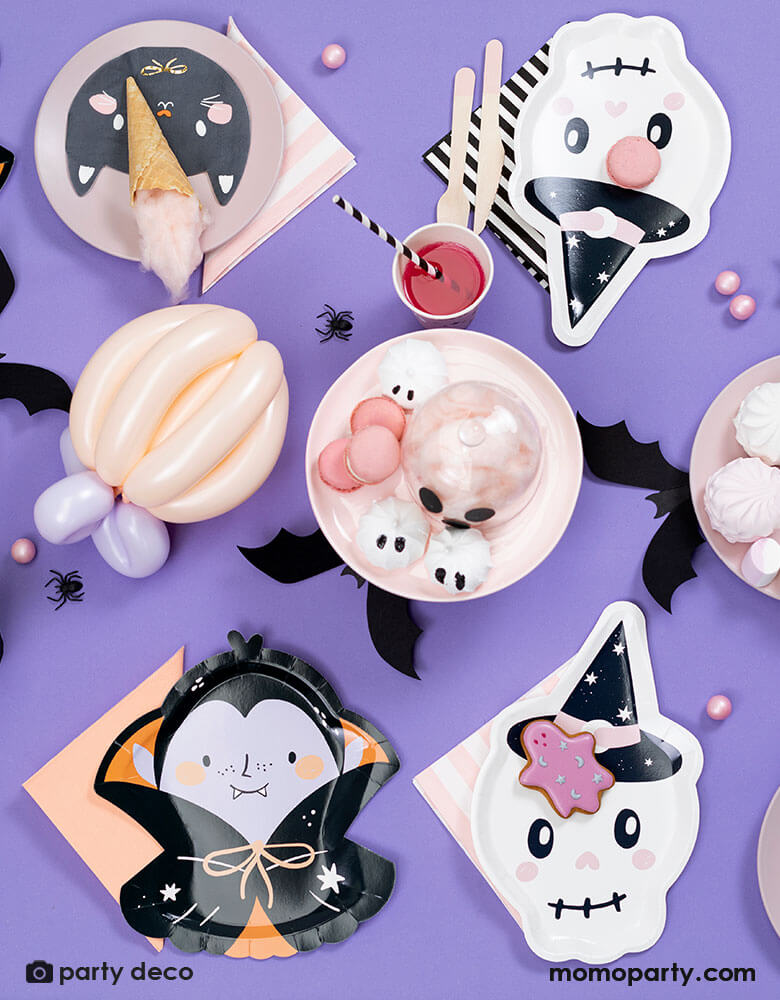 A purple Halloween party table featuring 7" x 10.6" pink skull shaped plates, Dracula shaped plates and black cat shaped napkin and pink striped napkin from Momopartys. Around the tableware there are some pink Halloween themed treats including marsh mellows, macaroons, cotton candies, with bat and spider confetti scattered around the table, it makes a great inspiration for a kid-friendly Halloween party.