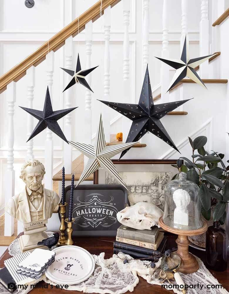 A table next to a staircase decorated with Momopartys's Salem Apothecary themed Halloween decorations and party goods including multiple black and white 3D hanging stars, Halloween black and white vintage themed bamboo tray and black and white vintage inspired paper plates and napkins. On the table there are also table statue and spell books as table decorations, making it a spooky yet elegant Halloween decoration inspiration.