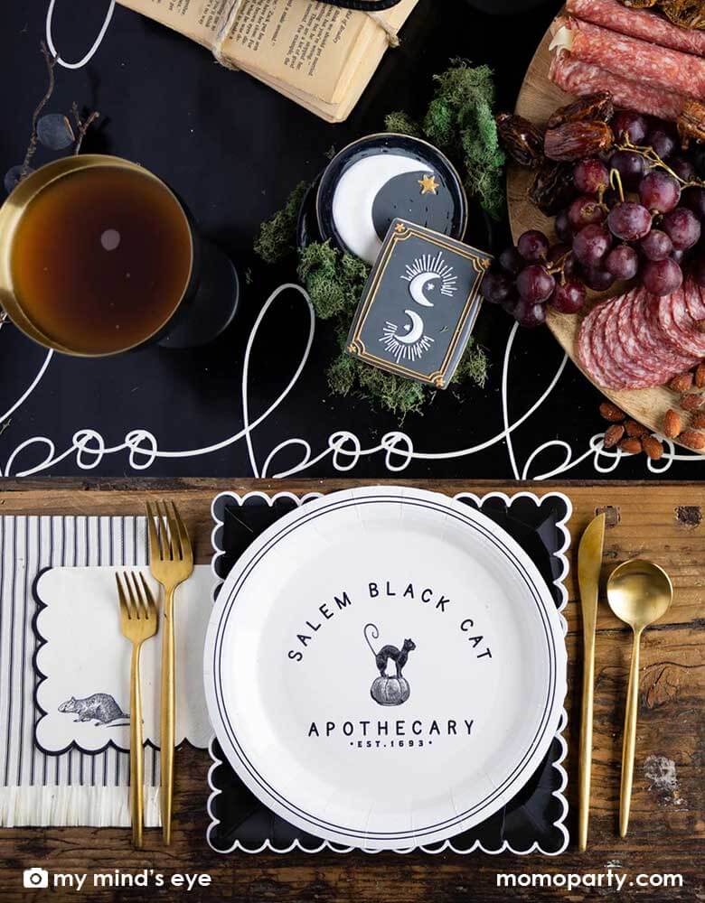 A elegant chic Halloween tablescape featuring Momopartys's Salem-Apothecary-Halloween-party supplies including a round plate, a black scalloped edged plate and Salem Apothecary Black Ticking Fringe Dinner Napkins by My Mind's Eye. On the table there is Salem Apothecary Boo Table Runner with Halloween themed crescent moon sugar cookies and a charcuterie board board. Makes it a great inspo for an elegant Halloween table.