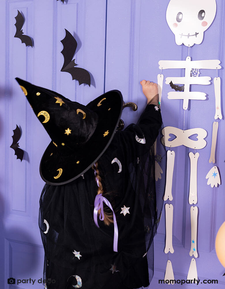 A little girl in witch costume knocking on a purple door for Halloween trick or treat. The door is decorated with Momopartys's skeleton papery decoration and paper bat decorations on it. A spooky fun vibe is in the air this Halloween season!