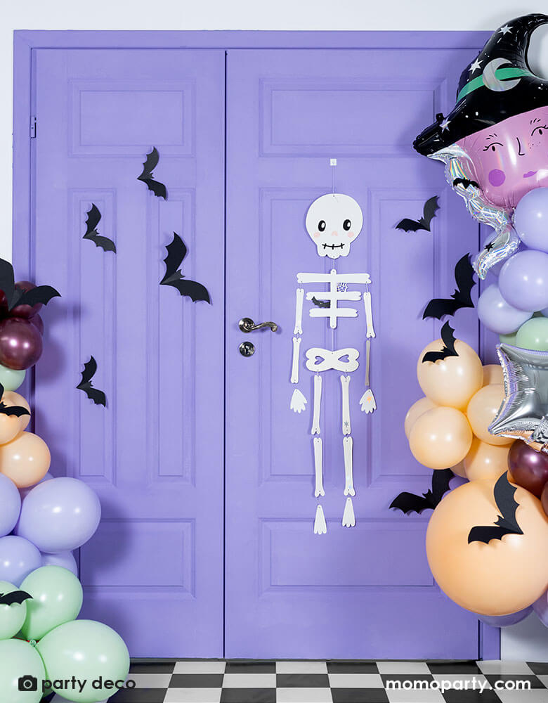 A purple door decorated with Halloween themed balloons and decorations including Momopartys's skeleton hanging paper decoration, witch head shaped foil balloon and a Halloween themed balloon garland in lilac, mint, blush and maroon colors, around the balloons there are paper bat decorations to create a spooky vibe a fun Halloween party.