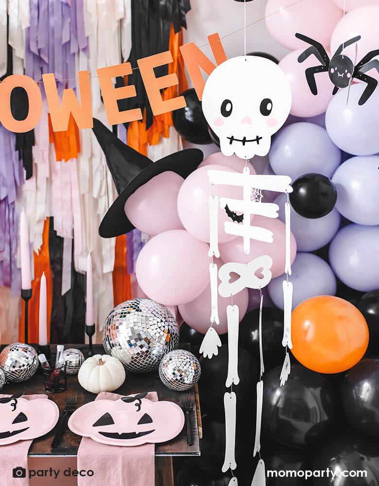 A festive Halloween party set up featuring a beautiful balloon garland in orange, pink, lilac and black, with Momopartys's 14" orange round skull foil balloon, witch's hats, skeleton hanging decoration, paper spider hanging decoration and honeycomb bats in front of a wall of colorful fringe in the Halloween themed colors. In the front there's a party table with some honeycomb pumpkins, disco balls and pink pumpkin shaped plates. All makes a great insp for a kid friendly Halloween bash.