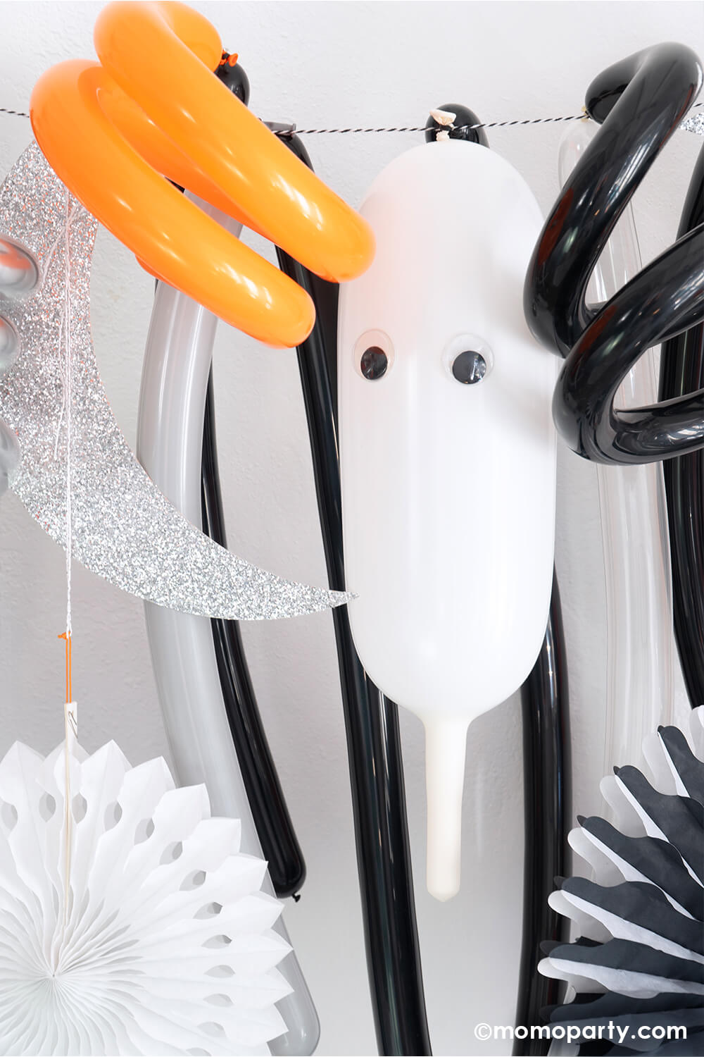 A fun whimsy Halloween themed long balloon backdrop by Momopartys featuring curl and long balloons decorated with googly eyes, handing honeycomb decorations and party fans in the classic Halloween colors of black, white and orange.