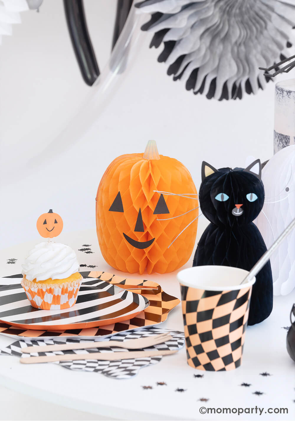 A fun Halloween party table in the classic black and orange colors featuring Momopartys's Halloween Checker Plates, napkins and party Cups by Meri Meri. On the table spider shaped confetti is scattered with honeycomb cat and pumpkin paper decorations as the centerpiece. On the plates there's a cupcake topped with a Halloween pumpkin topper. All makes a fun and whimsy inspiration for kid's friendly Halloween party ideas.