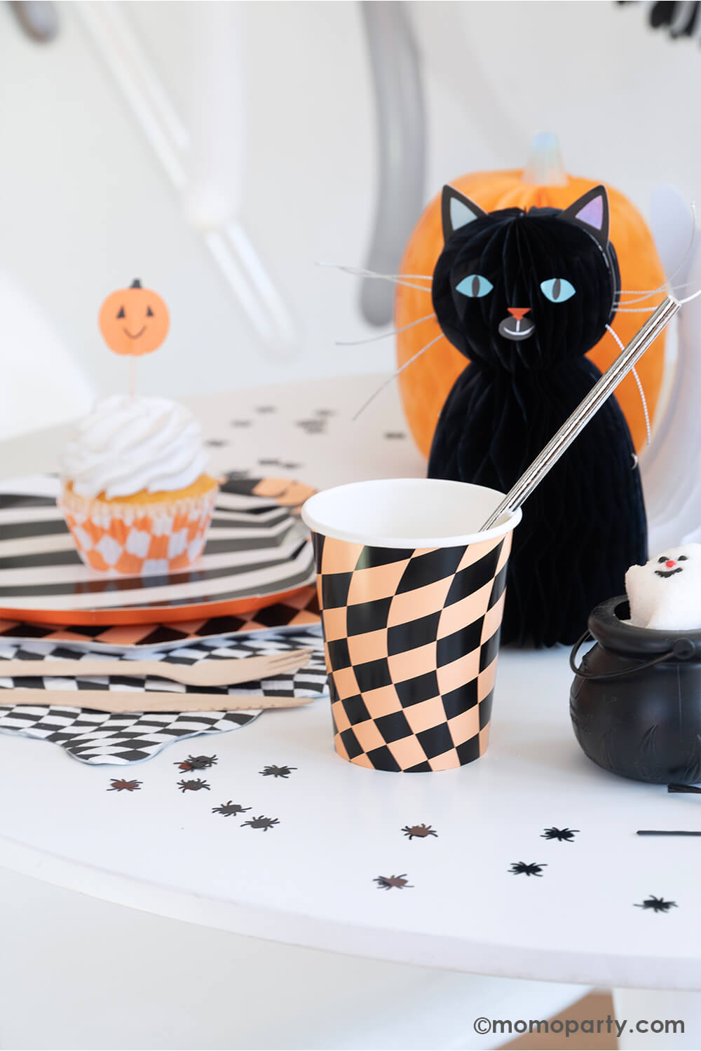 A fun Halloween party table in the classic black and orange colors featuring Momopartys's Halloween Checker Plates, napkins and party Cups by Meri Meri. On the table spider shaped confetti is scattered with honeycomb cat and pumpkin paper decorations as the centerpiece. At the corner there's a mini cauldron container filled with ghost shaped marshmallow treats. All makes a fun and whimsy inspiration for kid's friendly Halloween party ideas.