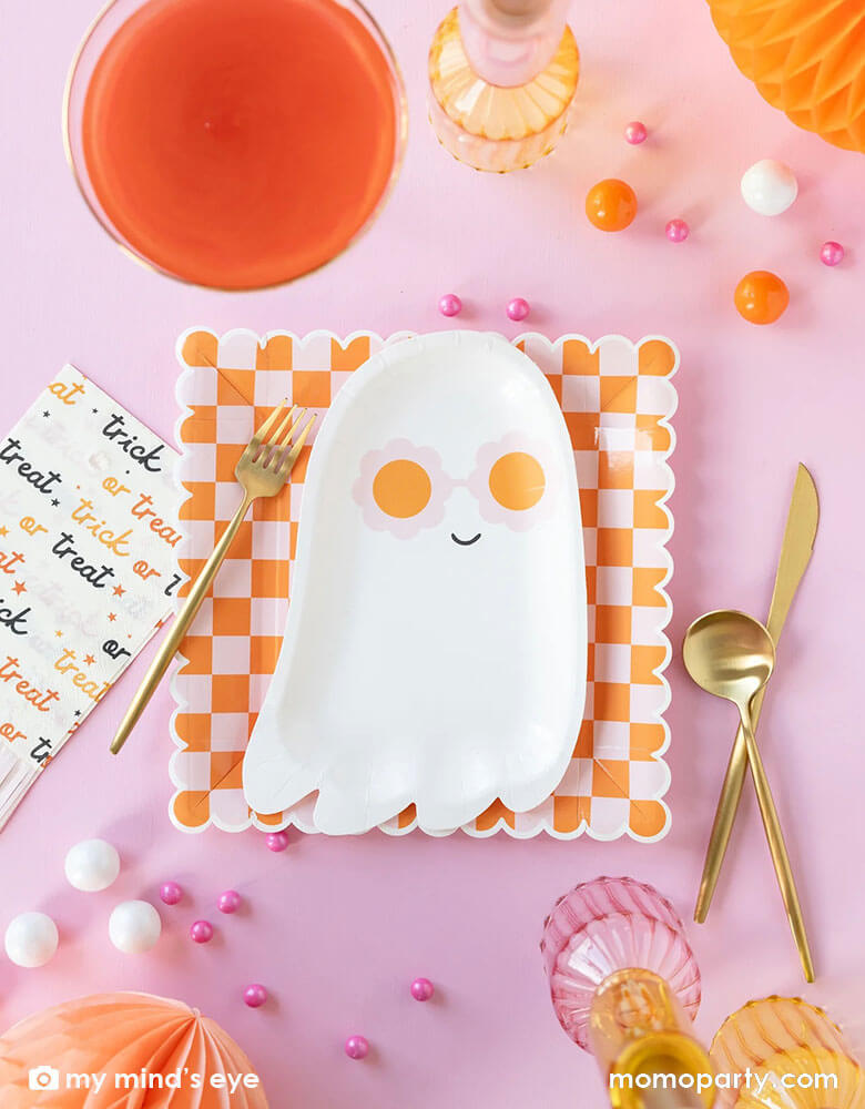 A pink Halloween table featuring Momopartys's pink sunny ghost shaped plates paired with orange/pink checkered plates, with pink trick or treat fringe napkins and honeycomb pumpkins around, it gives a great inspiration for a modern and chic Halloween party table decoration ideas.