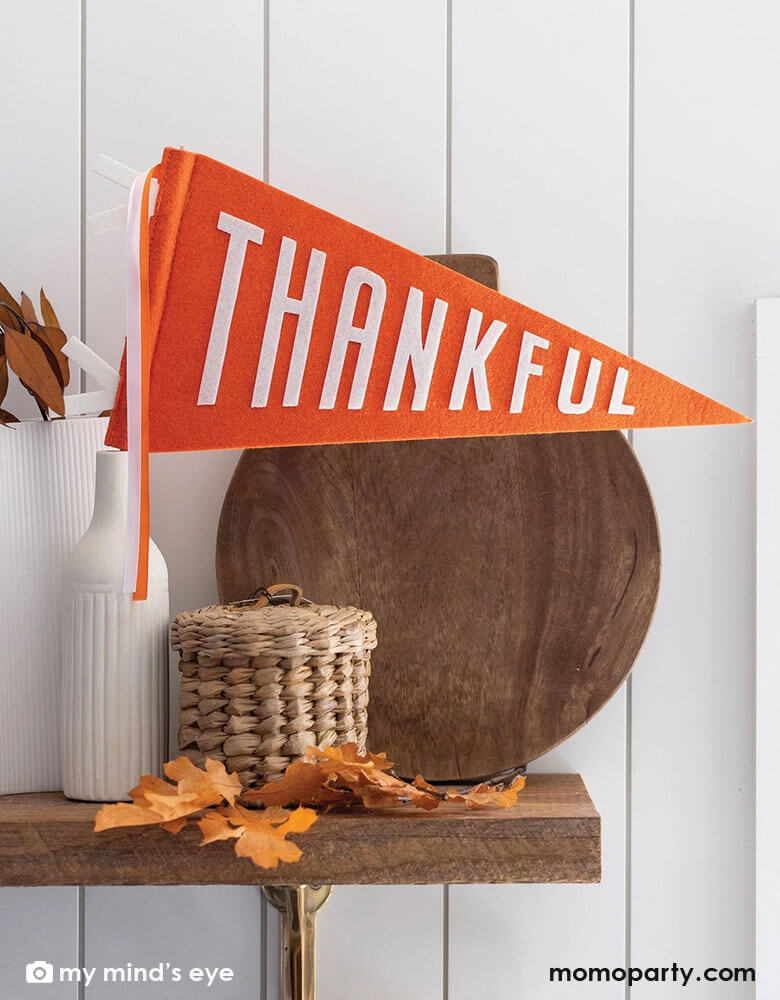 A fall/autumn wooden shelf decorated with a wooden cutting board, a weaved jar, some fall leaves and a couple white vase which one is adorned with Momopartys's 14" orange Thankful felt party pennant by My Mind's Eye. 