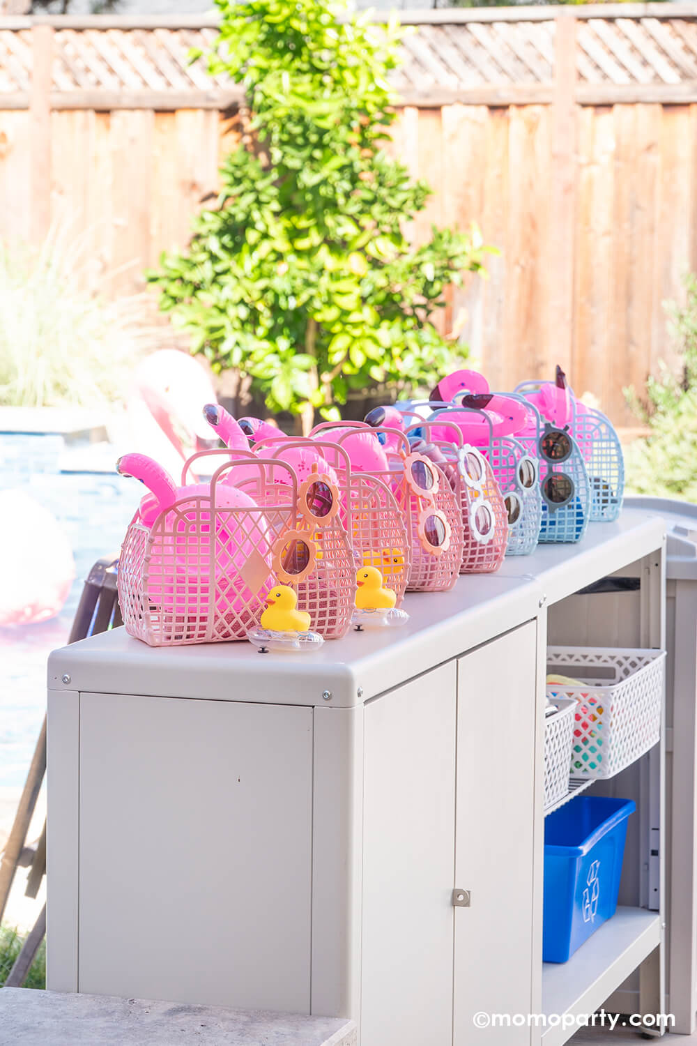 A bunch of vintage inspired jelly tote bags in pastel pink and blue lined up on pool side cabinet in a kid's pool party. These jelly tote bags are used as kid's take home goodie bags which have flamingo toys, beach towels, kids sunglasses and rubber duck toys. This adorable and fun goodie bag idea is perfect for a fun summer pool, beach or water themed celebration in the sun!