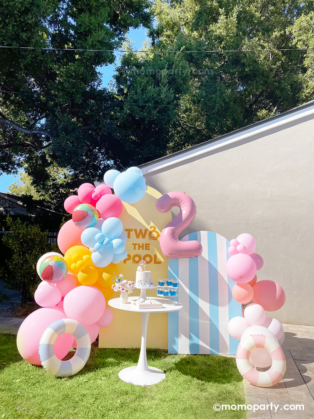A bright and pool inspired balloon installation for kid's 'Two The Pool' Second Birthday Party by Momopartys featuring latex balloons in pastel pink, yellow and blue adorned with beach ball foil balloons and vintage inspired pastel swimming ring inflatables in pastel blue and pink, with cabana inspired striped backdrop and a dessert table, this is a perfect set up for a summer-y sunny celebration!