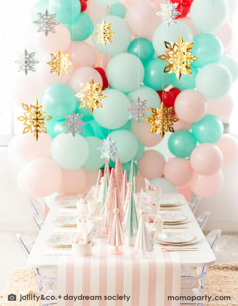 A pink Christmas party table featuring Momopartys's magical unicorn plates and cups by Daydream Society. A few ceramic Christmas tree decorations in mint and pink are used as the centerpiece on a pink white striped table runner. In the back of the table there's a balloon wall with pastel pink and mint balloons adorned with gold and silver snowflake hanging decorations, all makes it a whimsical kid's Christmas party inspo. 