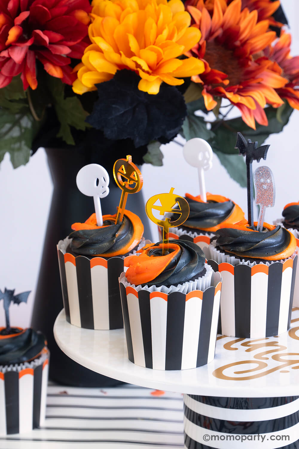 A close-up shot of Halloween orange and black swirled cupcakes in Momopartys's vintage Halloween black striped food cups topped with monster mash acrylic toppers featuring Halloween characters like pumpkin, skull, bat and ghost. The cupcakes are sitting on a black striped cupcake holder which is placed on a black striped table runner. On the table there's a fall flower arrangement with creepy black leaves, making this a perfect inspo for a fun and festive Halloween bash in this spooky season.